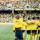 Soccer players in yellow jerseys celebrate a goal on the field as fans cheer in the stands, with one player raising his arms in victory.