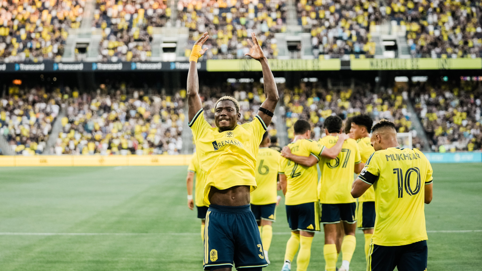 Soccer players in yellow jerseys celebrate a goal on the field as fans cheer in the stands, with one player raising his arms in victory.