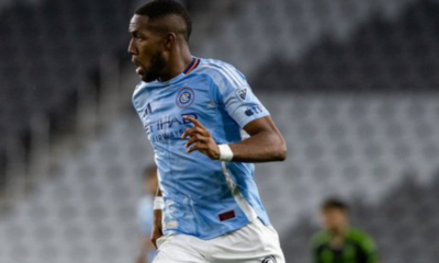 Soccer player in a light blue Manchester City jersey sprinting on the field during a match, with a blurred crowd in the background.