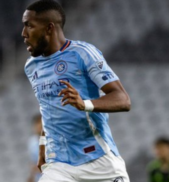 Soccer player in a light blue Manchester City jersey sprinting on the field during a match, with a blurred crowd in the background.