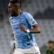 Soccer player in a light blue Manchester City jersey sprinting on the field during a match, with a blurred crowd in the background.