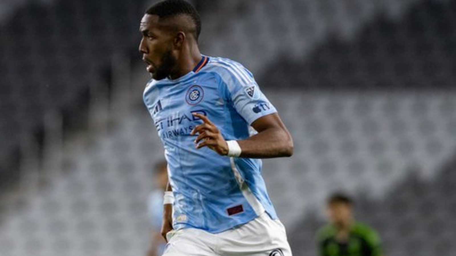 Soccer player in a light blue Manchester City jersey sprinting on the field during a match, with a blurred crowd in the background.