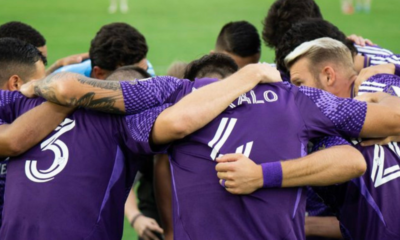 Soccer players in purple jerseys form a close huddle on the field, arms around each other in a team embrace.