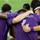 Soccer players in purple jerseys form a close huddle on the field, arms around each other in a team embrace.