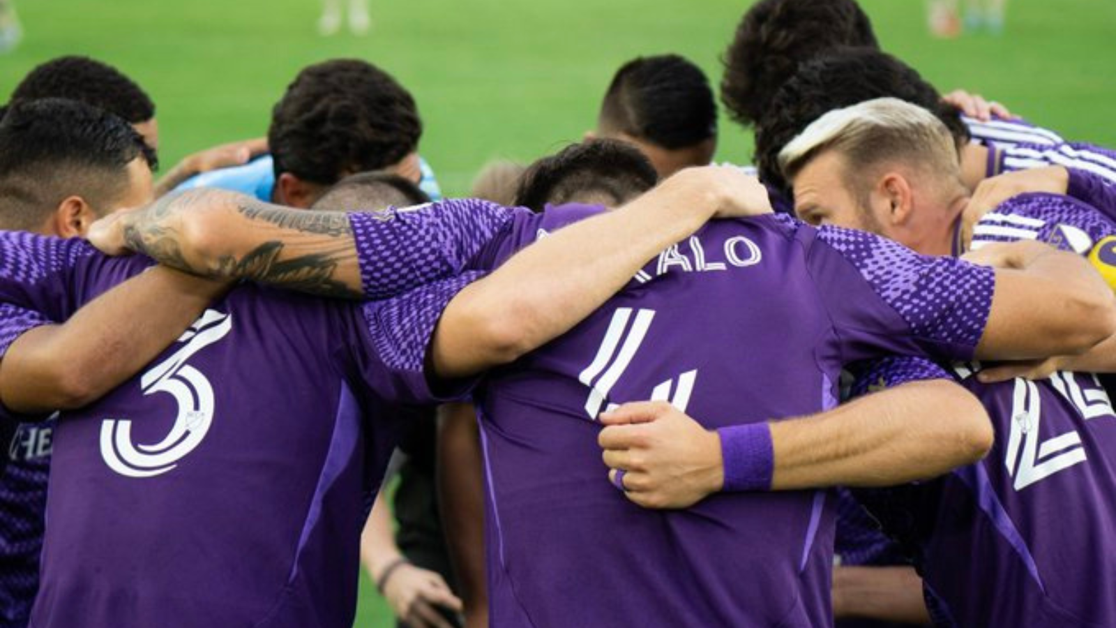 Soccer players in purple jerseys form a close huddle on the field, arms around each other in a team embrace.