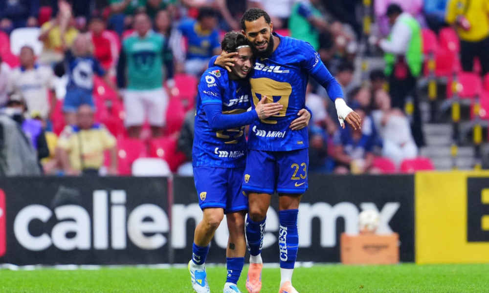 Two soccer players in blue uniforms hug on the field amid cheering fans in colorful seats, celebrating a goal.