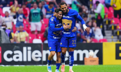 Two soccer players in blue uniforms hug on the field amid cheering fans in colorful seats, celebrating a goal.