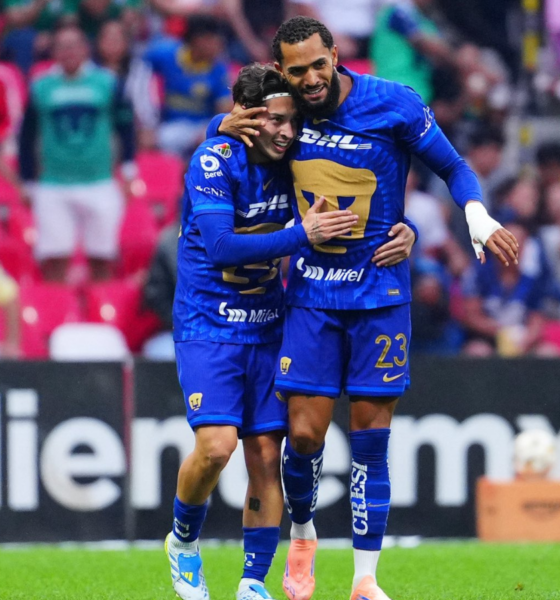 Two soccer players in blue uniforms hug on the field amid cheering fans in colorful seats, celebrating a goal.
