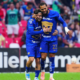 Two soccer players in blue uniforms hug on the field amid cheering fans in colorful seats, celebrating a goal.