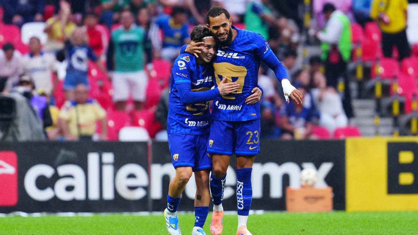 Two soccer players in blue uniforms hug on the field amid cheering fans in colorful seats, celebrating a goal.