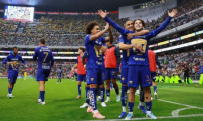 Soccer players in blue jerseys celebrate a goal on the field as fans cheer in the stadium stands.
