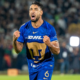 Soccer player in a blue DHL jersey celebrating with clenched fists on the pitch, crowd in the stands behind him.