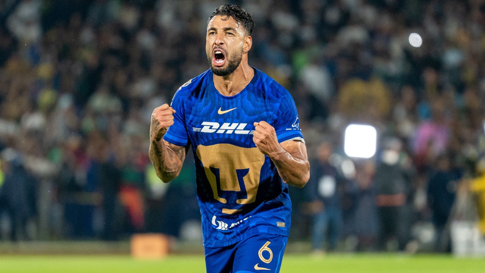 Soccer player in a blue DHL jersey celebrating with clenched fists on the pitch, crowd in the stands behind him.
