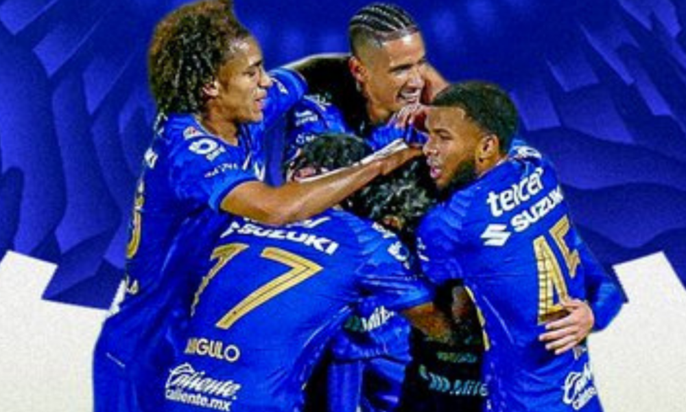 A group of blue-clad soccer players celebrate, embracing in a huddle with smiles after a goal.