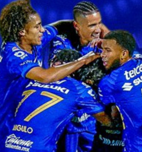 A group of blue-clad soccer players celebrate, embracing in a huddle with smiles after a goal.