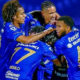 A group of blue-clad soccer players celebrate, embracing in a huddle with smiles after a goal.