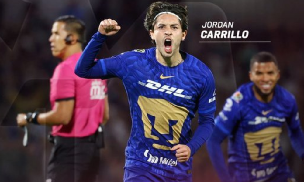 Soccer player Jordan Carrillo in blue jersey celebrates a goal, fist raised; teammate behind and a pink referee shirt visible left.