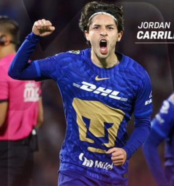 Soccer player Jordan Carrillo in blue jersey celebrates a goal, fist raised; teammate behind and a pink referee shirt visible left.
