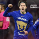 Soccer player Jordan Carrillo in blue jersey celebrates a goal, fist raised; teammate behind and a pink referee shirt visible left.