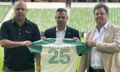 Three men stand on a football field, holding a green and white jersey with the number 25 in front of them on a sunny day.