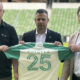Three men stand on a football field, holding a green and white jersey with the number 25 in front of them on a sunny day.