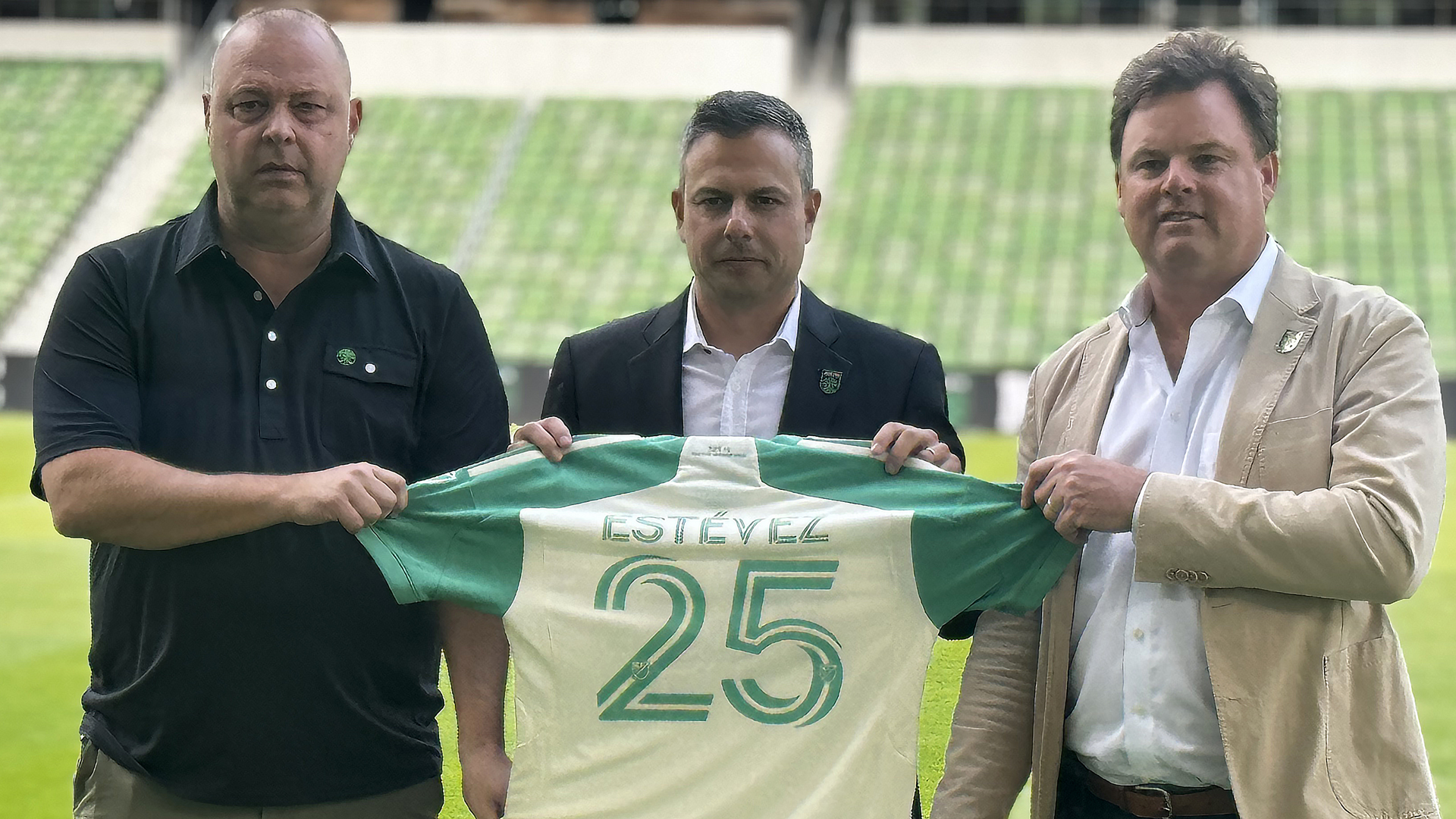 Three men stand on a football field, holding a green and white jersey with the number 25 in front of them on a sunny day.