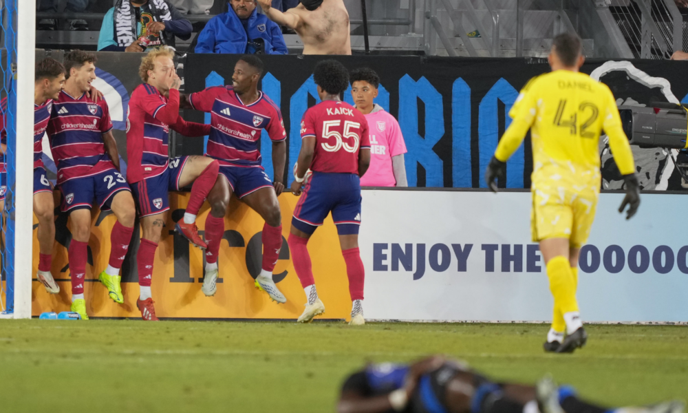 Soccer players in red and blue celebrate near the goal as a yellow-clad goalkeeper stands nearby in the foreground.