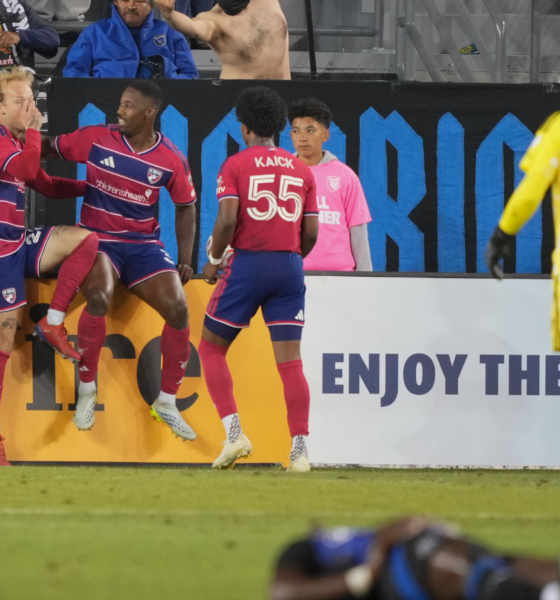 Soccer players in red and blue celebrate near the goal as a yellow-clad goalkeeper stands nearby in the foreground.