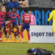 Soccer players in red and blue celebrate near the goal as a yellow-clad goalkeeper stands nearby in the foreground.