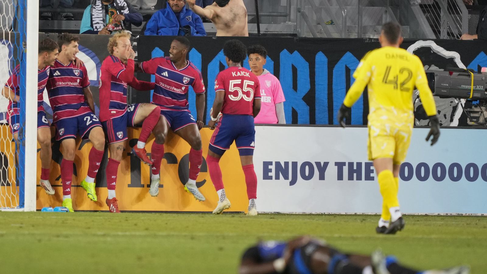 Soccer players in red and blue celebrate near the goal as a yellow-clad goalkeeper stands nearby in the foreground.