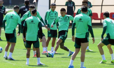 Soccer players in mint green training kits on a grassy field, forming a circle as one player kicks a ball during practice.