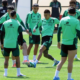 Soccer players in mint green training kits on a grassy field, forming a circle as one player kicks a ball during practice.