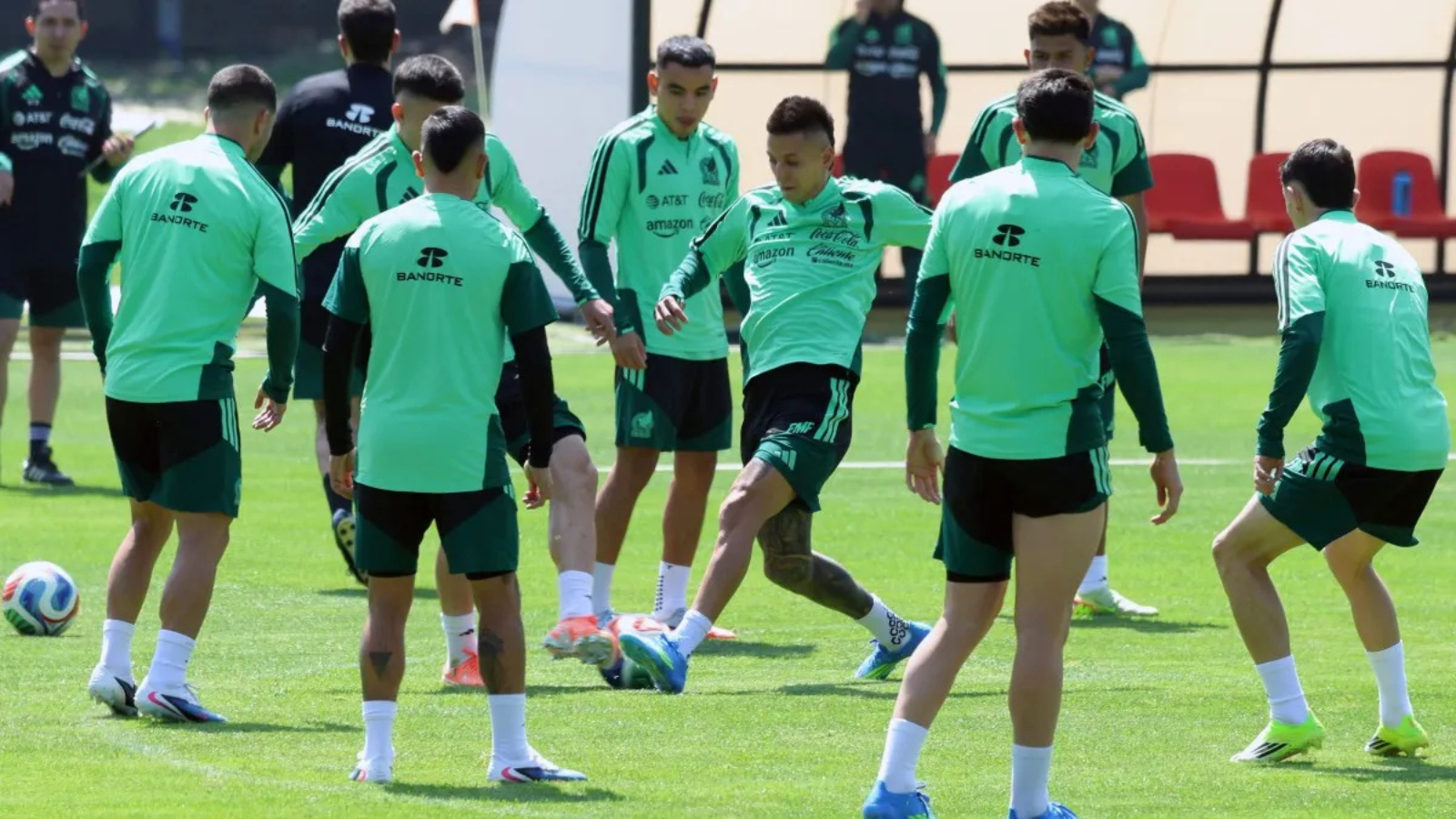 Soccer players in mint green training kits on a grassy field, forming a circle as one player kicks a ball during practice.