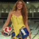 Smiling woman in a yellow sleeveless top and blue shorts holds a soccer ball in a stadium.