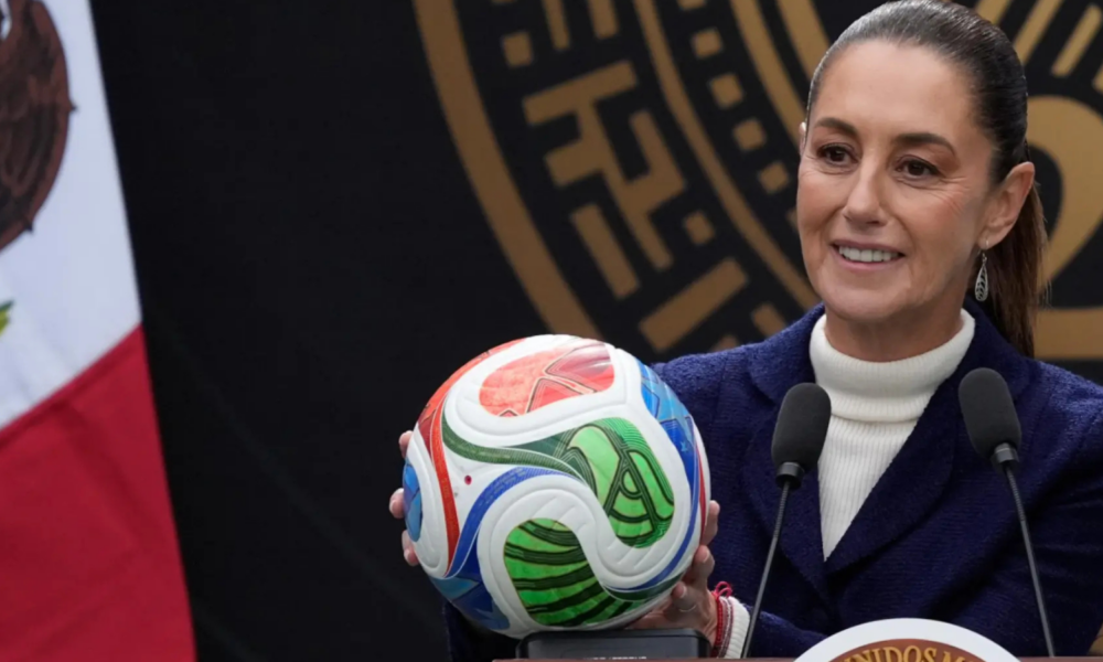 Smiling woman in a white turtleneck and navy blazer holds a colorful soccer ball at a podium with two microphones nearby, during a formal press event.