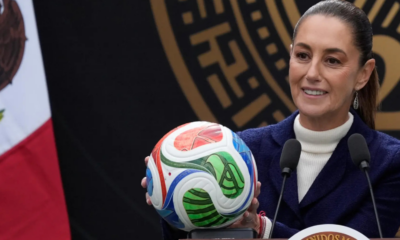 Smiling woman in a white turtleneck and navy blazer holds a colorful soccer ball at a podium with two microphones nearby, during a formal press event.