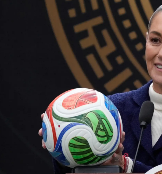 Smiling woman in a white turtleneck and navy blazer holds a colorful soccer ball at a podium with two microphones nearby, during a formal press event.