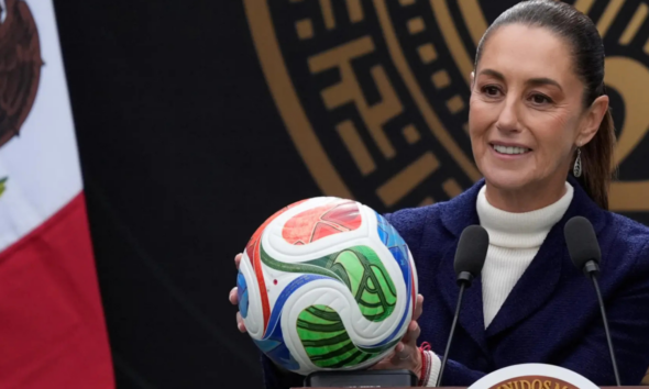 Smiling woman in a white turtleneck and navy blazer holds a colorful soccer ball at a podium with two microphones nearby, during a formal press event.