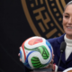Smiling woman in a white turtleneck and navy blazer holds a colorful soccer ball at a podium with two microphones nearby, during a formal press event.