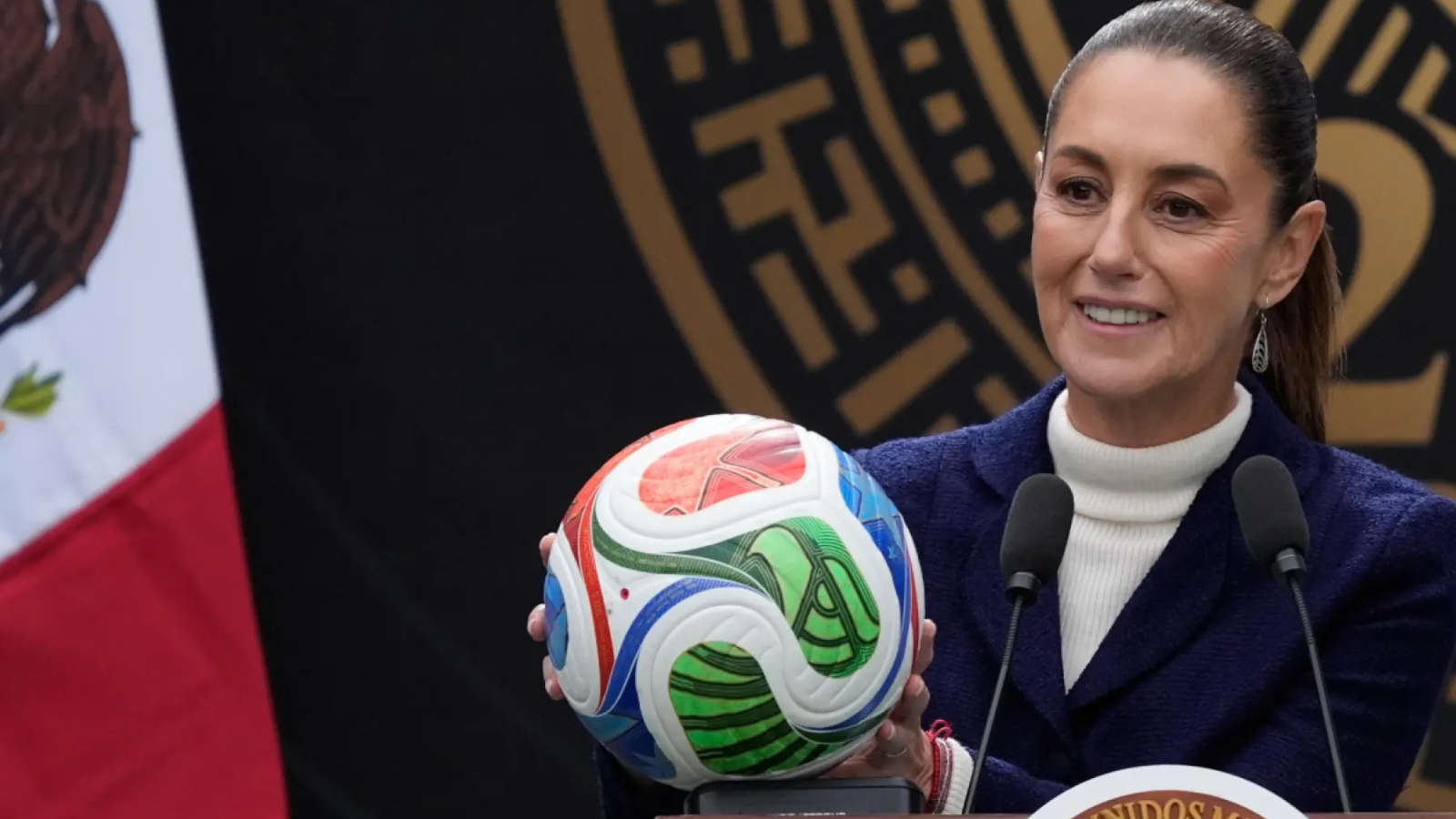 Smiling woman in a white turtleneck and navy blazer holds a colorful soccer ball at a podium with two microphones nearby, during a formal press event.