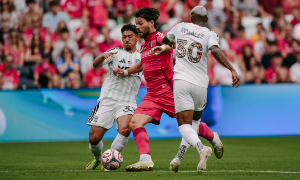 Soccer players contest the ball as a pink-clad attacker is boxed by two white-jersey defenders in front of a crowded stadium audience.
