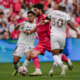 Soccer players contest the ball as a pink-clad attacker is boxed by two white-jersey defenders in front of a crowded stadium audience.