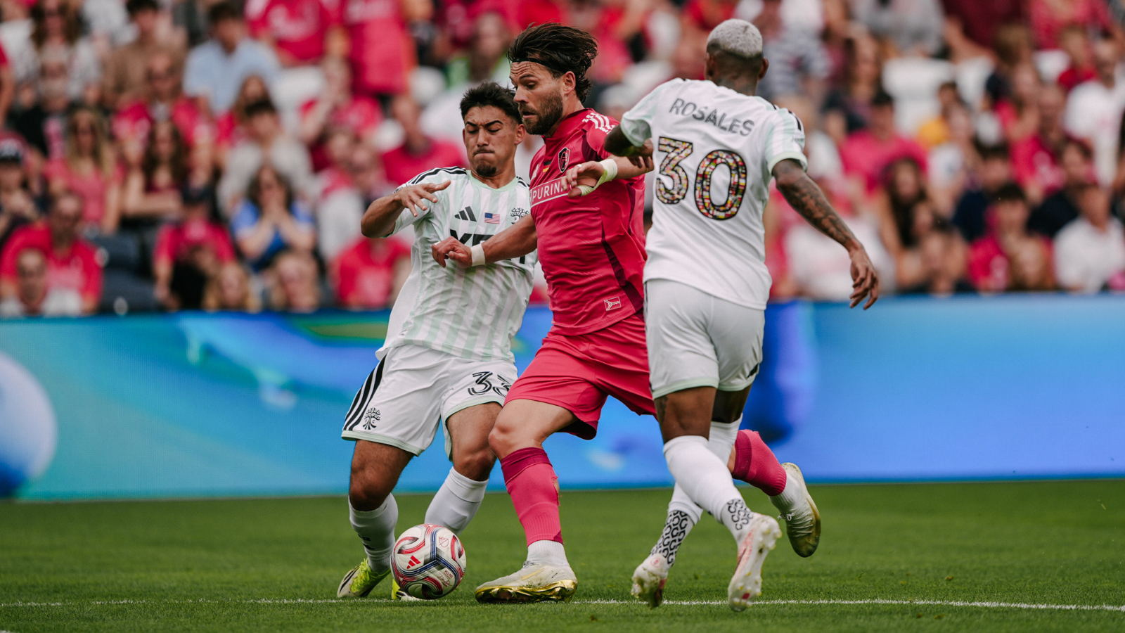 Soccer players contest the ball as a pink-clad attacker is boxed by two white-jersey defenders in front of a crowded stadium audience.