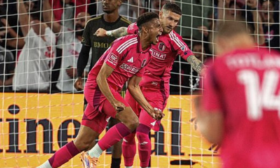 Soccer players in pink uniforms celebrate a goal near the goal net as fans cheer in the background.