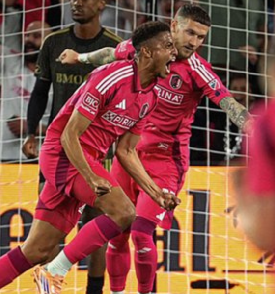 Soccer players in pink uniforms celebrate a goal near the goal net as fans cheer in the background.