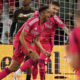 Soccer players in pink uniforms celebrate a goal near the goal net as fans cheer in the background.