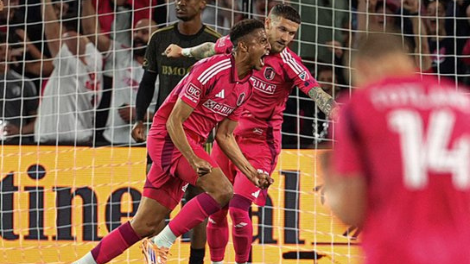 Soccer players in pink uniforms celebrate a goal near the goal net as fans cheer in the background.