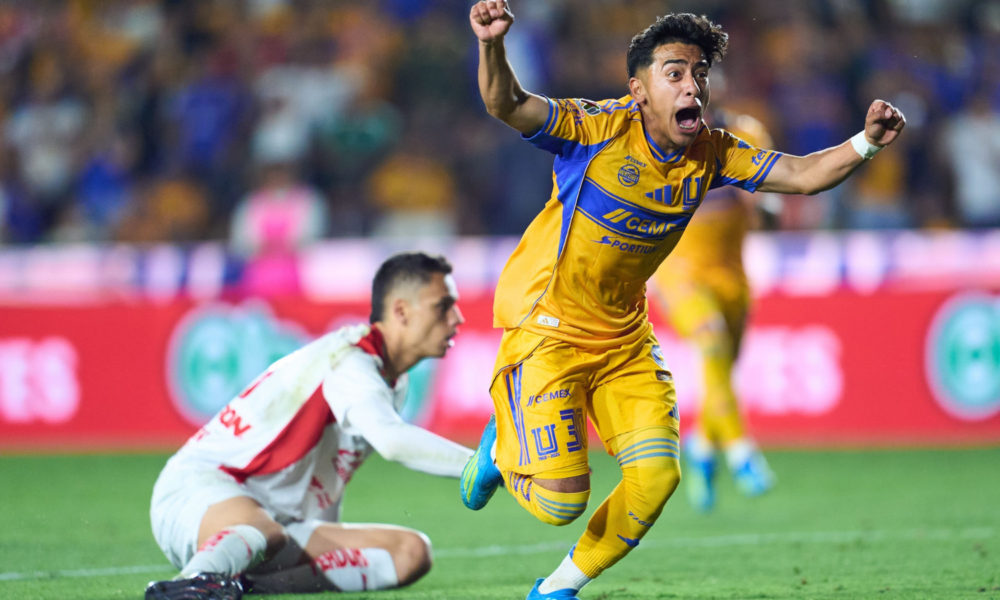 Soccer player in yellow kit jumps in celebration with arms raised, crowd in stadium behind him.