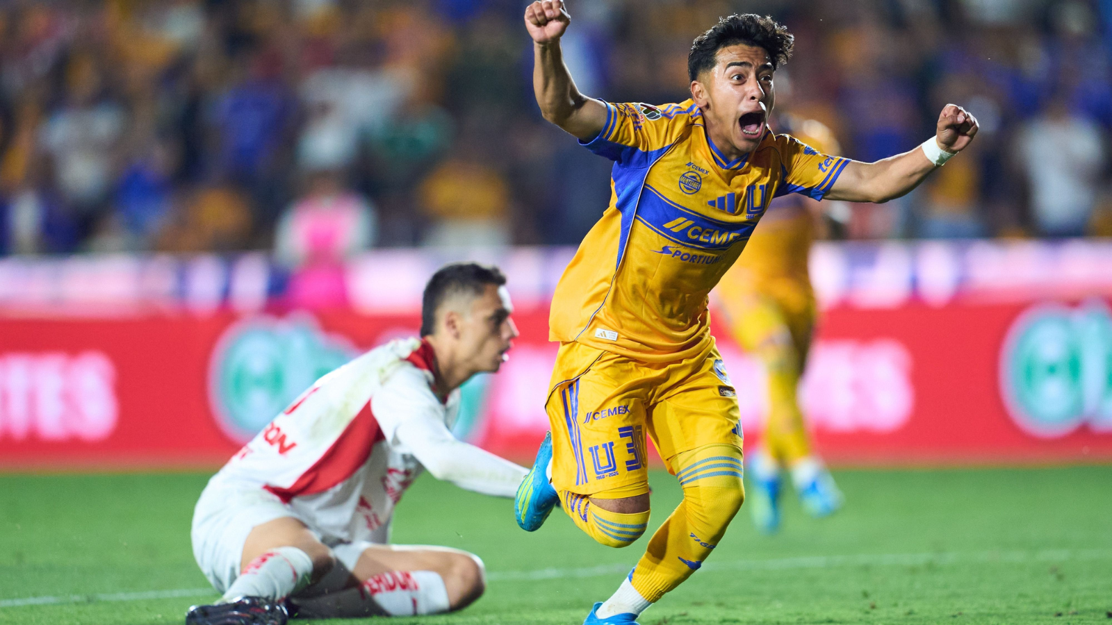 Soccer player in yellow kit jumps in celebration with arms raised, crowd in stadium behind him.