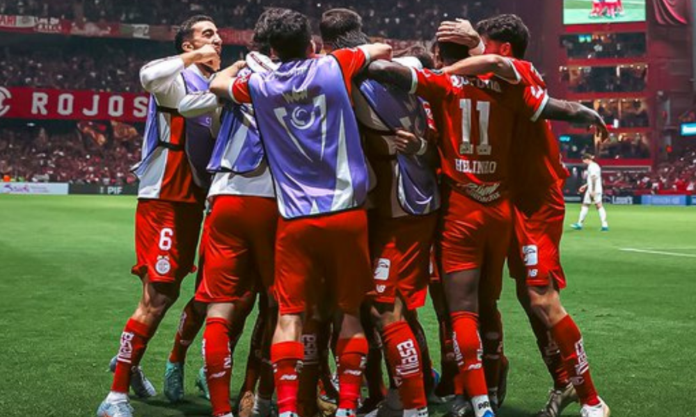Soccer players in red uniforms huddle together in celebration on a green field with a stadium crowd in the background.